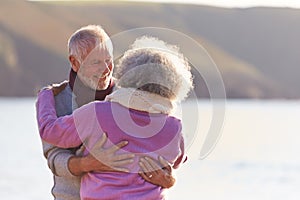 Loving Retired Couple Hugging Standing By Shore On Winter Beach Vacation Against Flaring Sun