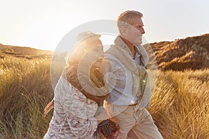 Loving Retired Couple Walking Arm In Arm Through Sand Dunes On Beach Vacation Against Flaring Sun