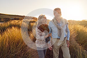 Loving Retired Couple Walking Arm In Arm Through Sand Dunes On Beach Vacation Against Flaring Sun