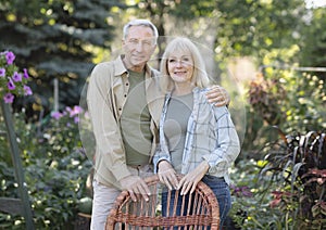 Loving retired spouses embracing, resting in garden in countryside, standing near wicker chair and smiling at camera