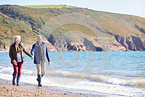 Loving Senior Couple Holding Hands As They Walk Along Shoreline Of Beach By Waves