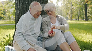 Loving senior couple hugging laughing having fun with apple during picnic in park