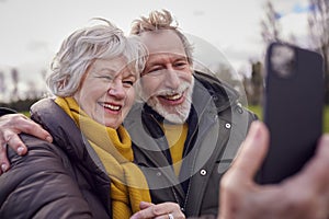 Loving Senior Couple Posing For Selfie On Mobile Phone On Autumn Or Winter Walk Through Park