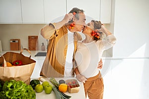 Loving senior spouses arriving from supermarket with grocery bag and unpacking, having fun and kissing in kitchen