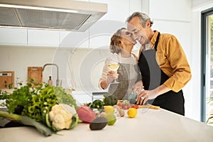 Loving senior spouses cooking together, woman kissing her husband, preparing dinner at cozy kitchen, free space