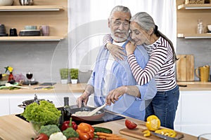 Loving Wife Embracing Her Elderly Husband While He Cooking Lunch In Kitchen