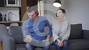 Loving woman talking to man as husband putting on headphones ignoring wife. Portrait of stressed sad Caucasian wife