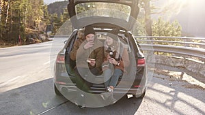 Loving, young couple drinks hot tea from thermos flask sitting in car trunk. Happy caucasian couple having a coffee