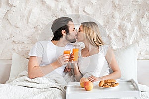 Loving young couple having breakfast in bed together, kissing, spending honeymoon at hotel