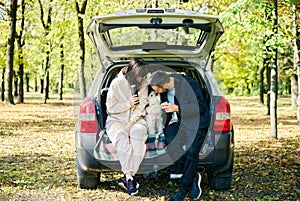 Loving young family petting and kissing his cute dog while sitting in car trunk relaxing in the forest