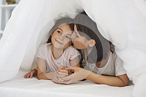 Loving young mother kisses her little daughter on the bed under the blanket in the bedroom.