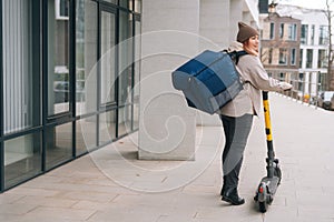 Low-angle back view of female courier food delivery with big thermal backpack walking with electric scooter in city