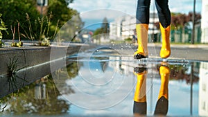 LOW ANGLE: Blurry woman in yellow rain boots runs into a big glassy puddle.