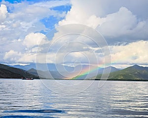 Low-angle of Como lake sunlit forested mountains rainbow cloudy sky background