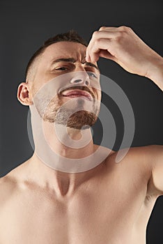 Low angle of concentrated frowning young bearded man plucking his eyebrows