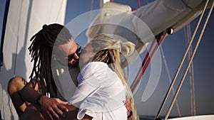 Low angle footage of stylish couple with dreadlocks in white clothes and sunglasses sitting embracing on the bow of the