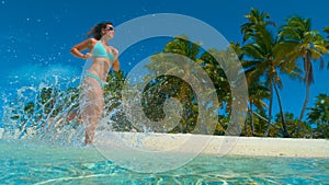 LOW ANGLE: Glassy ocean water splashes around girl running along exotic beach.