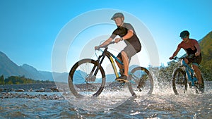 LOW ANGLE: Guys riding ebikes along the shallow river and splashing water