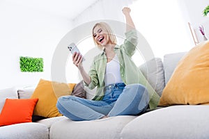 Low angle photo of crazy young woman sitting soft gray couch raise fist up with smartphone playing betting gambling