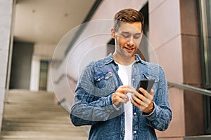 Low-angle portrait of happy handsome young man using mobile phone standing on stairs of modern office building