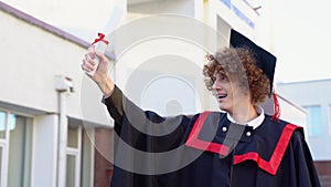 Low angle portrait of happy triumphant male graduate standing near university holding up diploma. From below of young