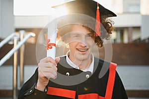 Low angle portrait of happy triumphant male graduate standing near university holding up diploma. From below of young