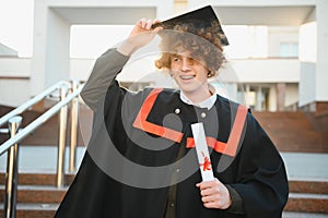 Low angle portrait of happy triumphant male graduate standing near university holding up diploma. From below of young