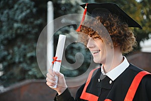 Low angle portrait of happy triumphant male graduate standing near university holding up diploma. From below of young