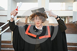 Low angle portrait of happy triumphant male graduate standing near university holding up diploma. From below of young