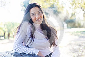 Low angle portrait of a smiling beautiful young hispanic woman sitting on park while looking camera