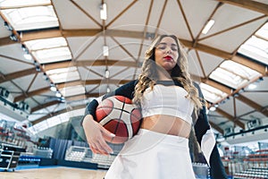 Low-angle shot of a blond cheerleader holding a basketball under her arm and squinting at the camera. Blurred bakground