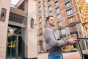 Low-angle shot of delivery man standing at city street near car and holding carton boxes with food.