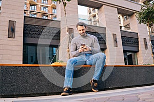 Low-angle shot of young man using mobile phone while sitting on urban bench at European city street.