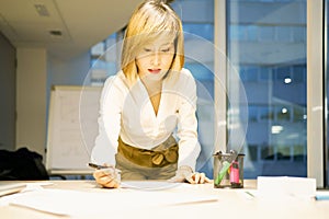 Low angle of smiling Asian businesswoman in elegant wear standing near table and reading paper reports while working in