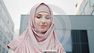 Low angle view of beautiful Muslim woman standing near modern office building with smartphone in hands. Young business