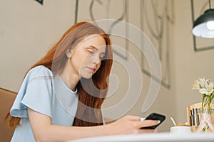 Low-angle view of charming young woman using smartphone texting message and drinking hot coffee sitting at desk in cozy