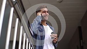 Low-angle view of cheerful young man talking on mobile phone holding coffee cup leaning on railing of office building.