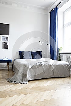 Low angle view of a cozy double bed in a corner of a bright bedroom interior with navy blue textiles and parquet floor