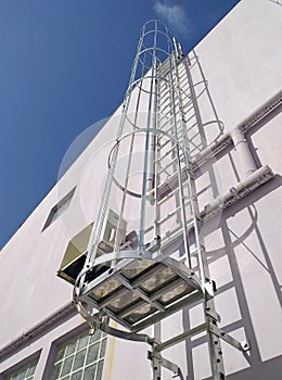Low angle view of an exterior vertical fixed ladder with safety cage in a building. White facade under blue sky. Building