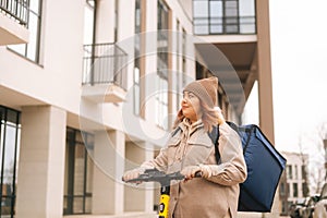 Low-angle view of female courier with big thermal backpack riding on electric scooter in city street looking away.