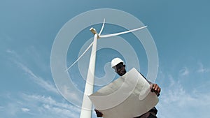 Low angle view of focused african american man standing on field with wind turbines and examining blueprints that