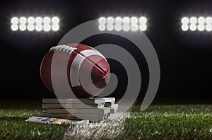 Low angle view of a football sitting on a pile of money on a grass field below stadium lights