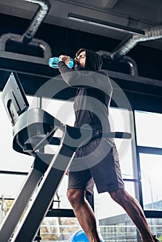 low angle view of handsome sportsman in hoodie exercising on treadmill and drinking water