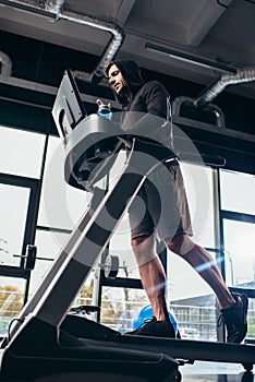 low angle view of handsome sportsman in hoodie running on treadmill and holding sport bottle with water photo