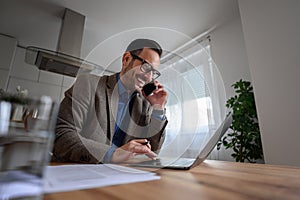 Low angle view of happy businessman talking on smart phone and working over laptop at home office