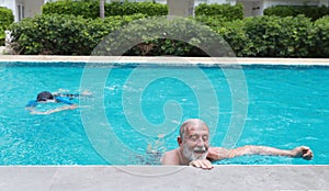 Low angle view happy elderly caucasian husband and elderly asian wife swimming in pool during retirement holiday with relaxation