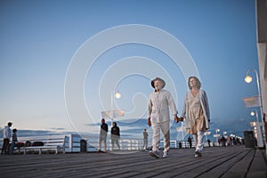 Low angle view of happy senior couple walking outdoors on pier by sea at dusk, holding hands.