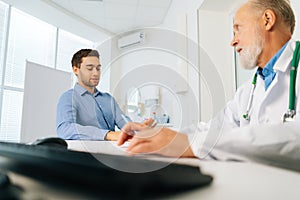 Low-angle view of male patient talking with mature adult doctor sitting at table during checkup visit in medical clinic