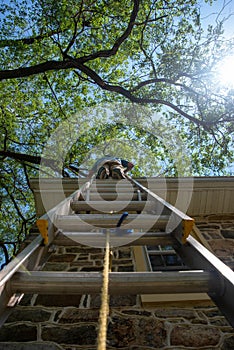 Low angle view of man on a ladder cleaning gutters of a stone house vertical image