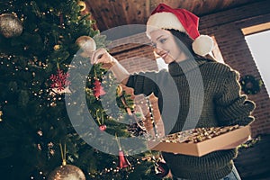 Low angle view photo of positive cheerful girl in santa claus red cap hold box with baubles hang christmas toy on fir